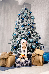 little girl child of five years sitting on the floor near Christmas tree decorated with toys, balls. In the hands holds an orange fruit tangerine, orange. dressed in a funny folder and a gray sweater