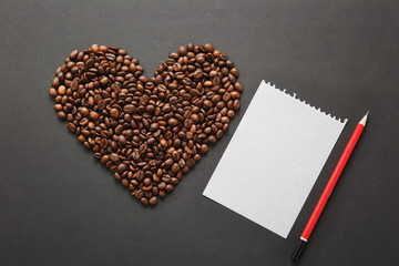 Brown coffee beans in form of heart isolated on black texture background for design. Saint Valentine's Day card on fabruary 14, holiday concept. Notebook, white blank sheet, red pencil. Place for text