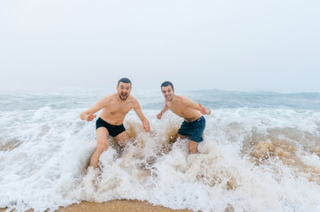 Two adult fellows screaming like crazy in cold water. Men friedns exressive faces after swimming in stormy scary ocean. Emotional male crazy faces fooling. Awesome fearless guys under wave strike.