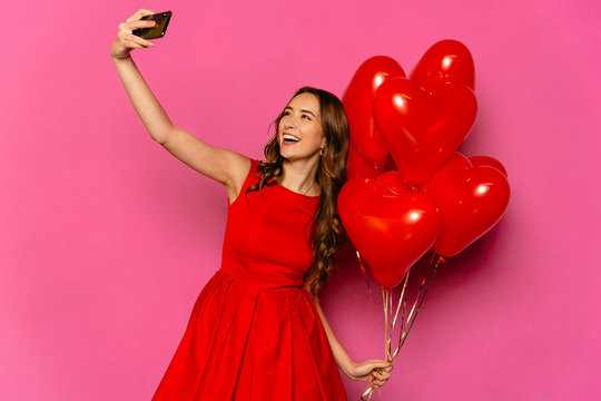 Young charming woman taking selfie on smartphone with heart shaped air balloons celebrating Valentine's day. Dressed in red dress.