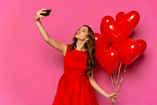 St. Valentine's day. beautiful woman taking selfie, giving a kiss, holding heart shaped air balloons. In red dress.