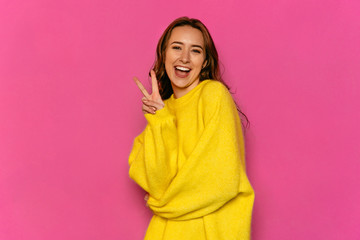 Gorgeous young woman showing peace gesture, dressed in yellow sweater. Over pink background. Concept of Valentine's day.