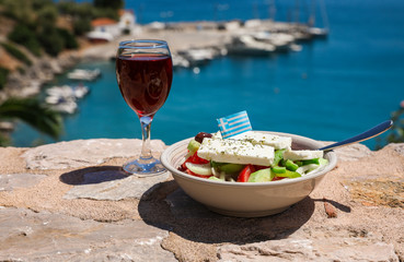 A glass of red wine and bowl of greek salad with greek flag on by the sea view, summer greek...