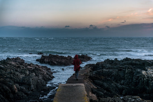 Young adult woman standing on a stone pier at the atlantic coast near a lighthouse in Ireland and watching the ocean and sunset