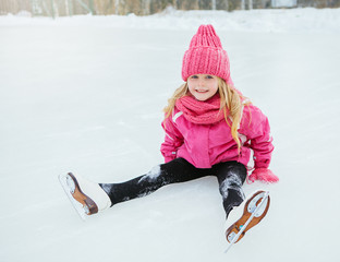 Little smiling girl skate and fell on ice in pink wear. Outdoor.