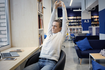 Picture of talented web designer sitting in front of open laptop pc and stretching his back, feeling sleepy and tired while working on big project. People, technology, job and occupation concept