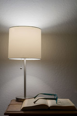 A pair of broken reader glasses resting on an old open book, underneath a bedside table lamp, against white wall, suggesting an elderly taking a break from reading  