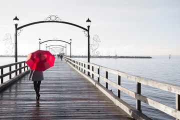 Woman holding a red umbrella walking on a rainy day on the pier in White Rock, British Columbia, Canada.