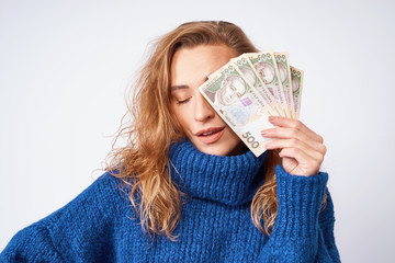 A cheerful woman in a sweater, covers part of the face with money hryvnia holding them in her hands on a gray background