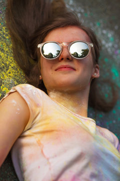 Closeup portrait of gorgeous model in mirror glasses lying on the asphalt covered paint Holi