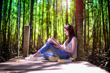 woman enjoy reading on wooden bridge under shadow and light of forest tree, feeling relax and comportatble easy listening music online from mobile device in the meanwhile