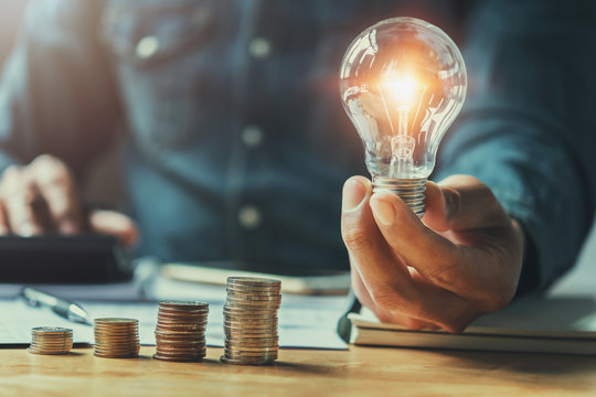 business man hand holding lightbulb with using calculator to calculate and money stack. idea saving energy and accounting finance in office concept