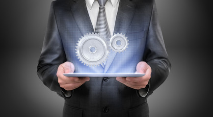 A businessman in a suit holds a glowing tablet with two interlocked metal spur gears hovering above...
