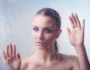 Young brunette woman with makeup smoky eyes has his hands on the glass with water drops closeup on a light background