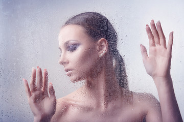 Young brunette woman with makeup smoky eyes leaned a hand against the glass with water drops closeup on a light background