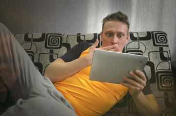 Fototapeta premium Frightened man lies on the couch and holds a tablet computer and watches a scary movie. Loafer.