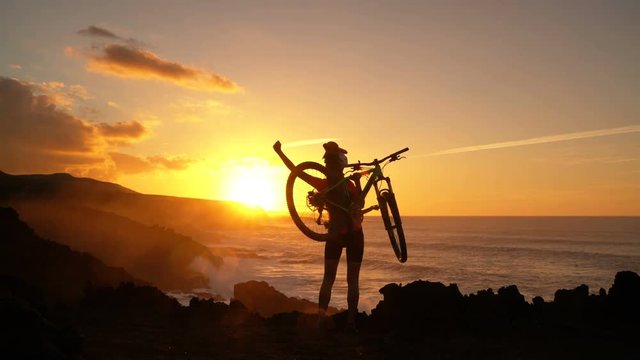 Aspirations, success and achievement. Active lifestyle MTB cyclist mountain biking woman cheering happy raising arms lifting bike by sea at sunset celebrating. Person with bicycle in amazing nature.