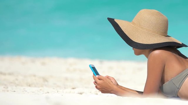 Woman using phone app on beach on summer travel vacation sunbathing on sand. Young female on smartphone lying on beach towel wearing sunhat by sea. She is wearing bikini while relaxing during summer.