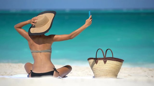 Stylish woman taking selfie on beach. Summer travel holidays concept while sunbathing on beach. Woman in bikini is photographing using smart phone on vacation.