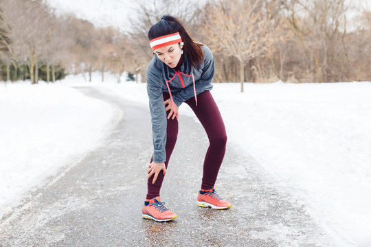 Portrait of Caucasian young woman feeling pain in legs after jogging outside in winter park. Girl having injury when running excercising  outdoors in cold weather conditions