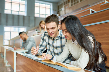 Obraz premium Portrait of two cheerful students sitting at desk in lecture hall of modern college, young man and Asian woman enjoying class, copy space