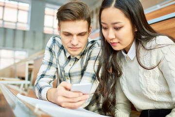 Portrait of two students, young man and Asian woman, using smartphone during break, sitting at desk in lecture hall of modern college, copy space
