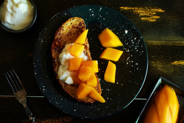 Top view of sweet french toast with yogurt and mango on black plate over dark background. 