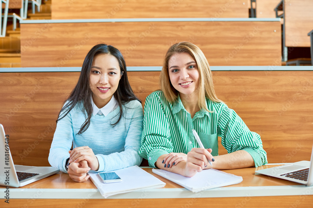 Wall mural portrait of two pretty girls in college, one of them asian, smiling ad looking at camera while sitti