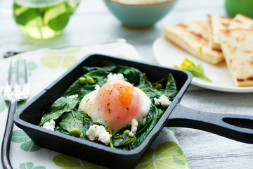 Closeup of poached egg with spinah, basil and feta in iron cast skillet with flatbread and green tea on background. Soft focus.