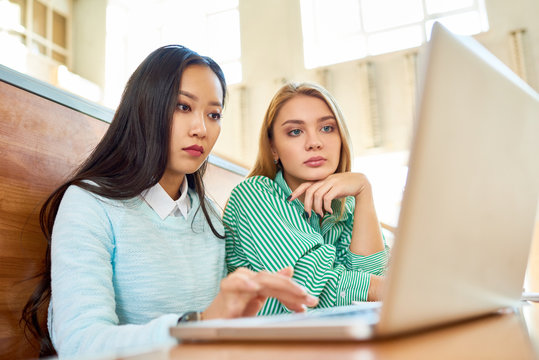 Portrait of two pretty girls in college, one of them Asian, sitting at desk in auditorium and using laptop