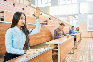 Portrait of beautiful Asian woman raising hand during class at college, copy space with people in background