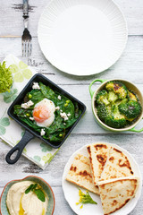Top view of green shakshuka with spinach, basil and feta in a pan, baked broccoli, hummus and pita bread, green mint tea on white table with empty plate. Oriental style breakfast or lunch.