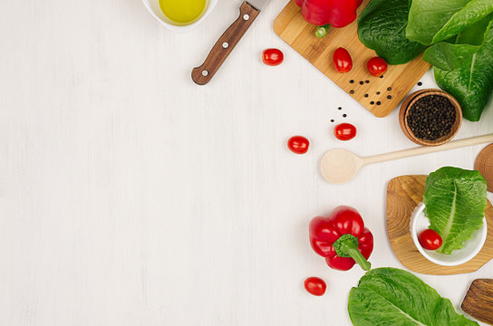 Border of fresh green greens, red paprica, cherry tomato, pepper, oil and utensils on soft white wooden background.