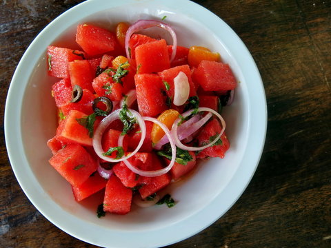 Cut watermelon spicy salad, ingredients with red onion, coriander, olive, olive oil, lemon, orange and chili on white plate top on wood table, copy space, summer menu concept 