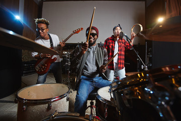 Band of young drummer, guitarist and singer playing instruments and performing songs in studio of...