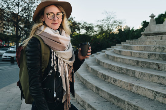 Sunny autumn day, backlight. Young attractive woman tourist in hat, eyeglasses and with backpack stands on city street,drinks coffee. Hipster girl walks, looks at sights. Vacation, adventure, trip.