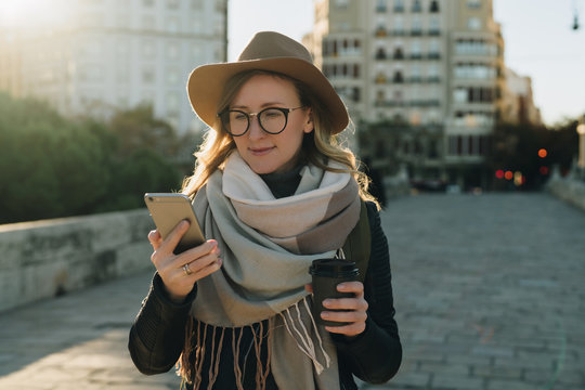 Sunny autumn day. Young attractive woman travels in hat and eyeglasses stands on city street, uses smartphone and drinks coffee. Hipster girl is looking for road. Vacation, adventure, trip.