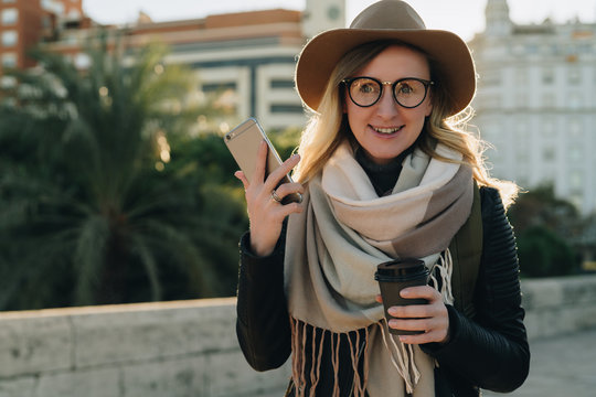 Sunny autumn day. Young attractive woman tourist in hat and eyeglasses stands on city street, uses smartphone and drinks coffee. Hipster girl is looking for road. Vacation, adventure, trip.