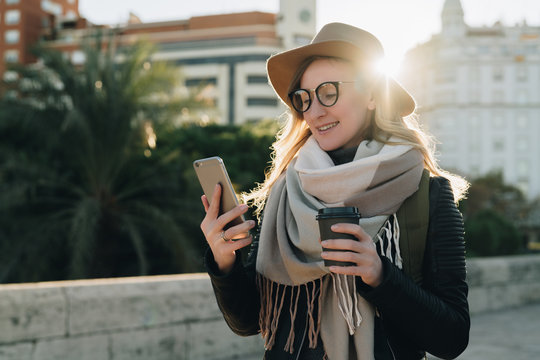 Sunny autumn day, backlight. Young attractive woman tourist in hat and eyeglasses stands on city street, uses smartphone and drinks coffee. Hipster girl is looking for road. Vacation, adventure.