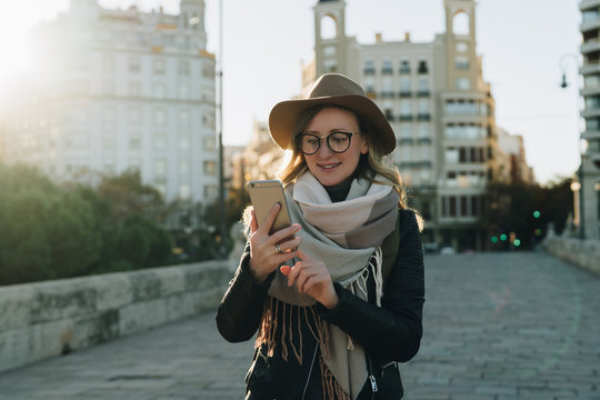 Sunny autumn day, backlight. Young attractive woman travels in hat and eyeglasses stands on city street, uses smartphone. Hipster girl is looking for road, sights. Vacation, adventure, trip.