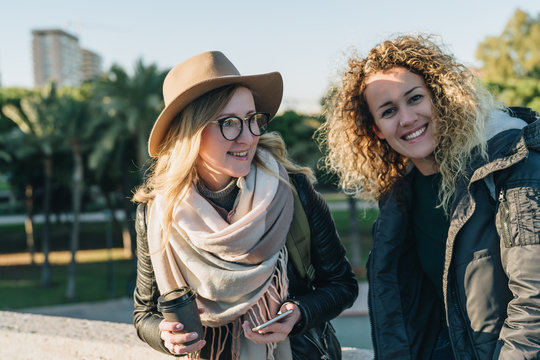 Sunny autumn day. Two young cheerful women tourists are standing on city street, drinking coffee. Girl in hat and eyeglasses is holding smartphone. Vacation, adventure, trip. Blurred background.