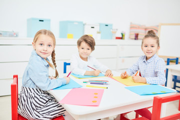 Cute fritndly kids in casualwear gathered by table in kindergarten to draw