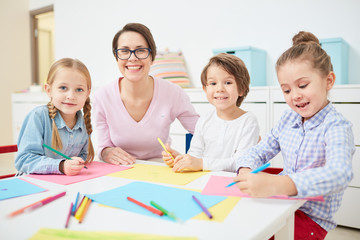 Happy young teacher and small group of adorable kids drawing with crayons in kindergarten