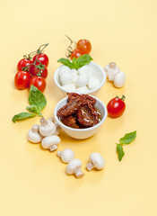 Sun dried tomatoes, champignon mushrooms, Mozzarella cheese and basil leaves on a light yellow background.