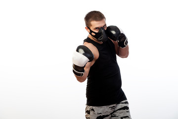 Young sporty blue-eyed man in sports black T-shirt, wearing boxing gloves and black training mask, boxing on white isolated background, front view