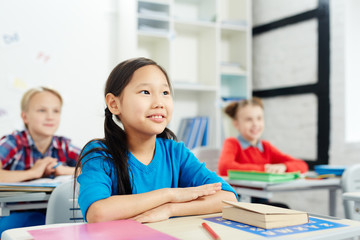 Fototapeta premium Little Asian schoolgirl sitting by desk with her classmates on background