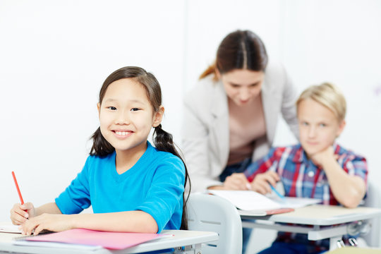 Youthful girl with crayon looking at camera with teacher and schoolboy talking on background