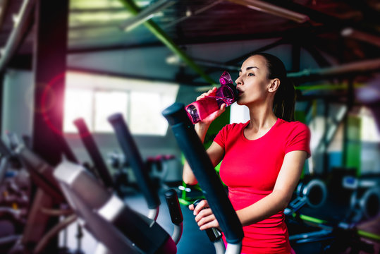 A short break to rest. An attractive young athlete in bright sports clothes is drinking water on a treadmill in the gym. Woman in gym