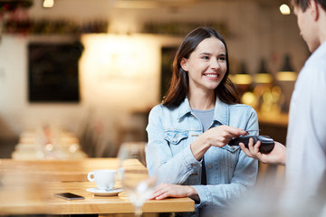 Young smiling woman with card holding it over payment terminal while paying for her order in cafe - obrazy, fototapety, plakaty