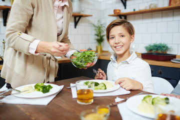 Naklejka premium Happy boy looking at camera by dinner while his grandmother putting him vegetable salad on plate